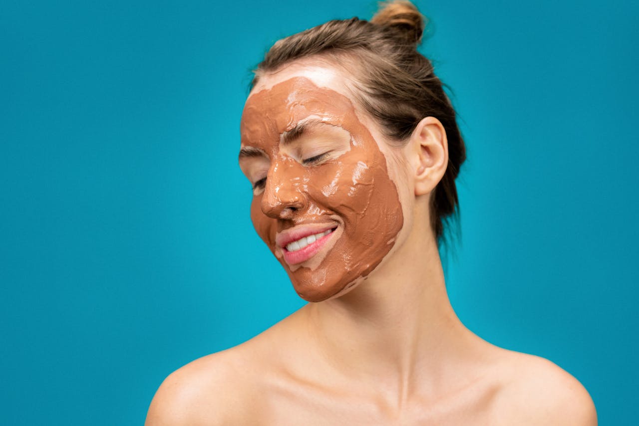 A woman smiles while applying a clay mud mask for skincare treatment.