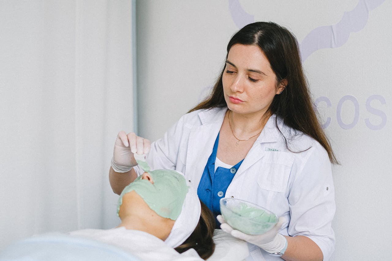 A beautician applies a green facial mask to a client in a wellness clinic, promoting skin care and relaxation.